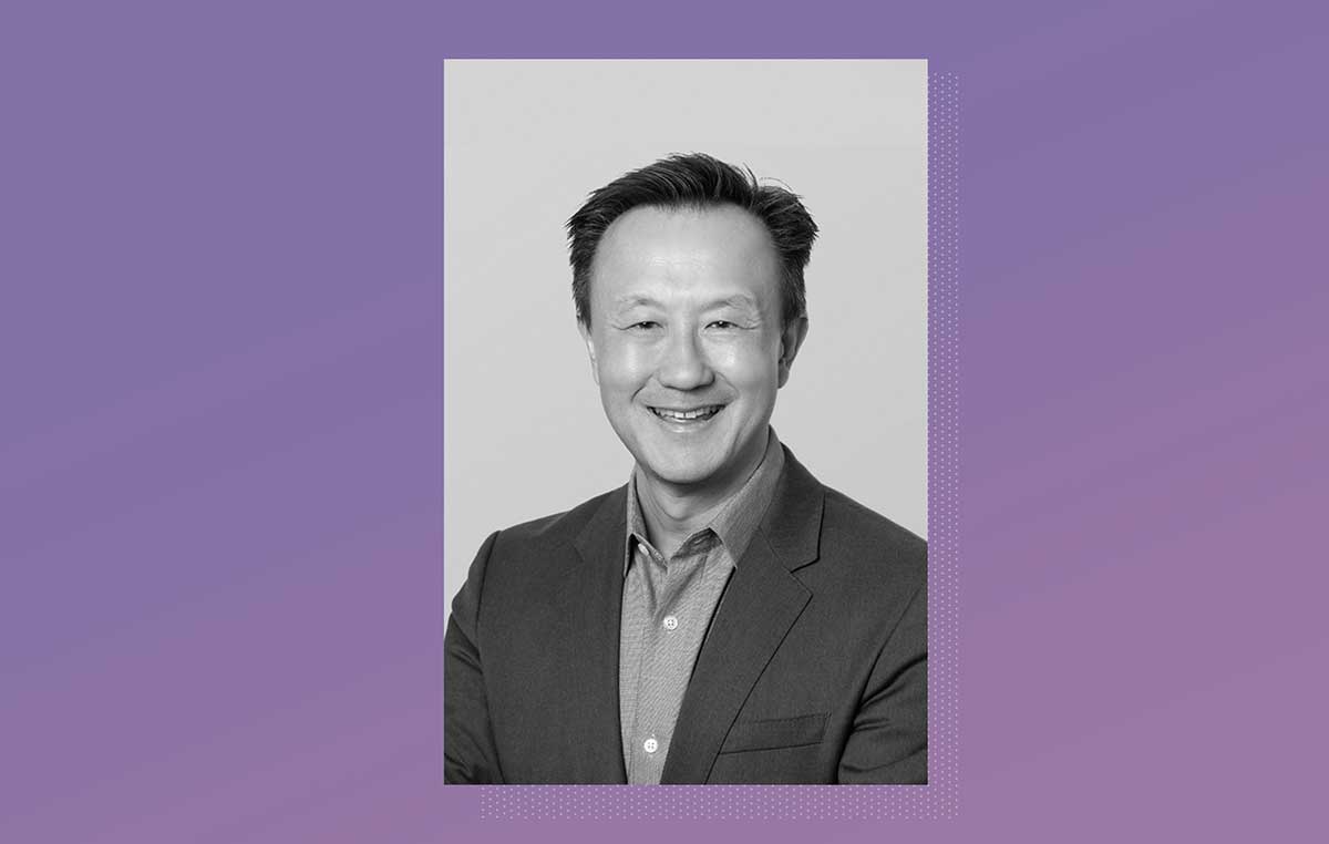 Grayscale photo of John Kuo, Chief Legal Officer for Visby Medical, in dark suit with no tie on light background, placed on a lavendar gradient backdrop.